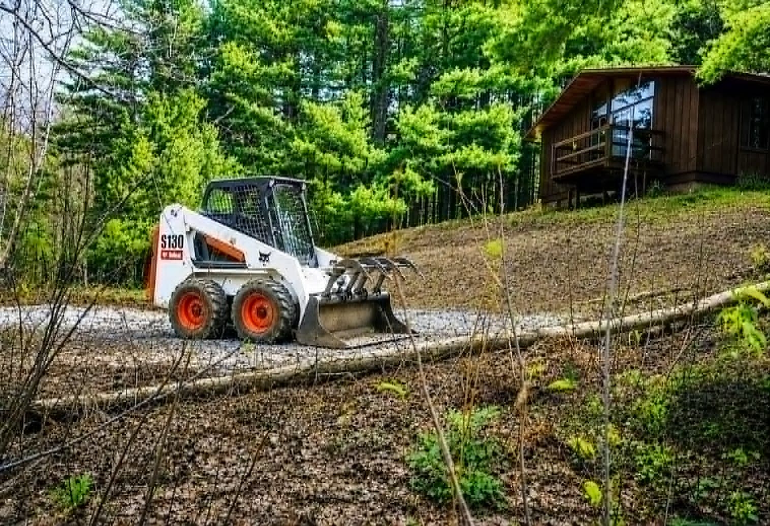 Bobcat loader with grapple attachment re-grading a sloped hillside near a home